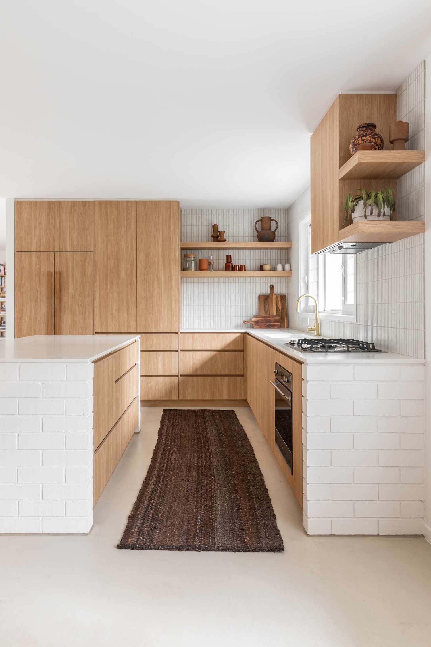 Modern-coastal-kitchen-featuring-warm-timber-cabinetry-brushed-brass-tapware-exposed-shelves-soft-concrete-flooring-and-white-brick-bench