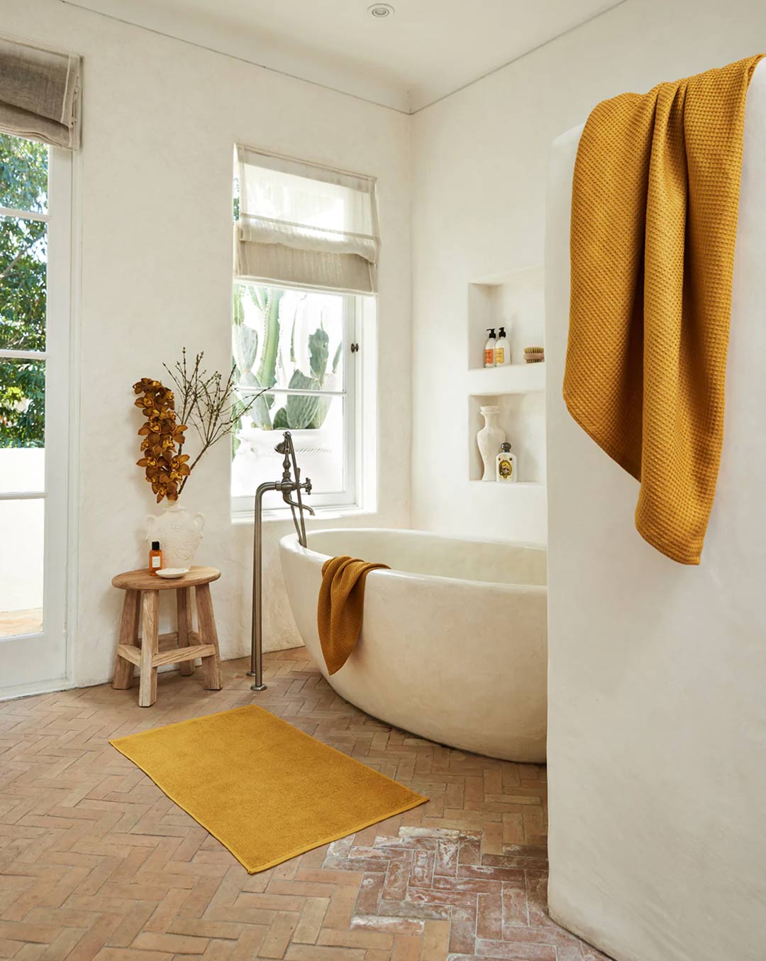 A-rendered-bathroom-with-a-concrete-bath-and-herringbone-brick-look-tiles-a-floor-mounted-bath-filler-and-mustard-yellow-towels-and-bath-matt