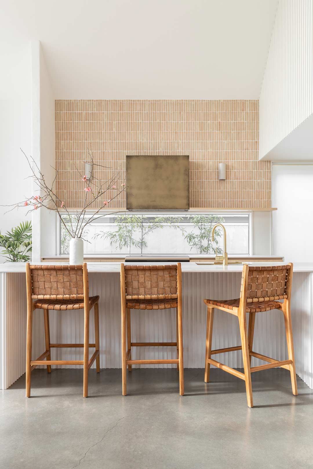 A-warm-open-kitchen-in-a-barn-style-house-with-large-island-bench-brushed-brass-tapware-clay-teracotta-tiles-and-woven-leather-bar-stools